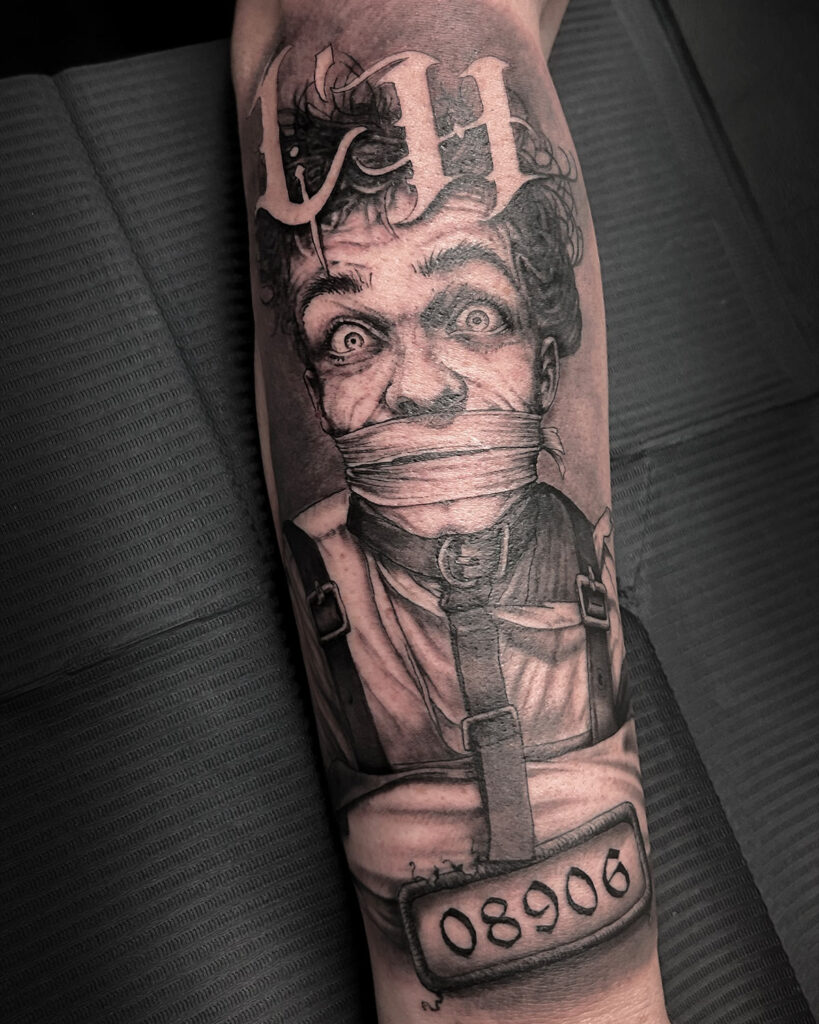 Tatuaje de un loco con camisa de fuerza, con las letras LH, un número, estilo realismo, en la zona del antebrazo, tatuado por kaorukel
